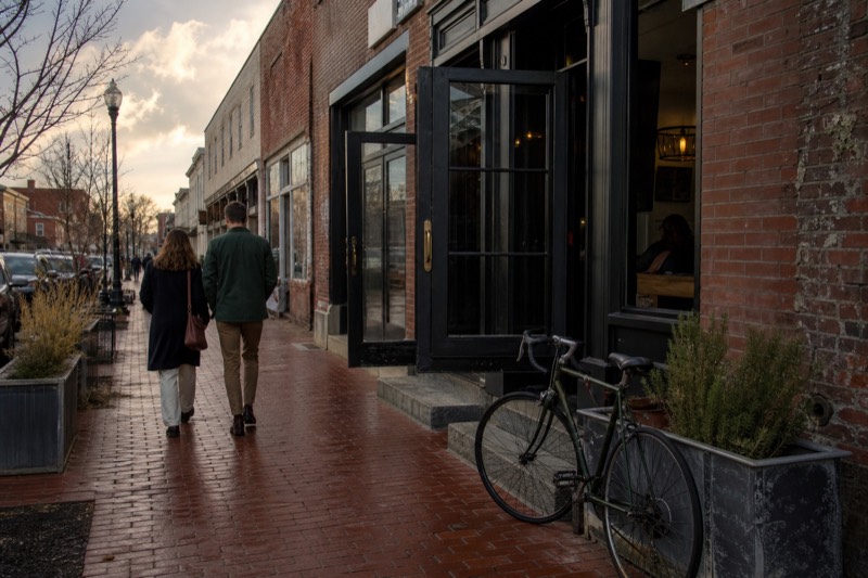 Brick storefronts line a sunny small-town street, bicycles at a rack, chalkboard outside a tasting room